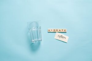 A minimalist composition of a glass with Scrabble tiles spelling 'HYDRATE' on a blue background.